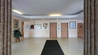 This building lobby offers a functional and welcoming entry point for residents and visitors alike. The space is defined by its light-toned large-format tile flooring, which extends throughout and is complemented by a dark entrance mat that guides guests inward. The ceiling features a distinctive recessed tray design with evenly spaced recessed lighting, creating a bright and clean atmosphere.

Two side-by-side elevator doors are prominently positioned along the back wall, providing vertical access throughout the building. Adjacent to the elevators, a bank of mailboxes is neatly mounted on the wall, offering a convenient mail retrieval area for residents. Framed informational boards and notices are displayed nearby, serving as a communication hub for building announcements.

Wall-mounted artwork adds a touch of character to the otherwise neutral, light-toned walls. The overall design is practical and straightforward, prioritizing ease of circulation and clear sightlines throughout the lobby. The space is well-maintained and efficiently laid out, making it easy to navigate upon entry. The combination of recessed lighting and light-colored surfaces ensures the lobby remains bright and inviting throughout the day.