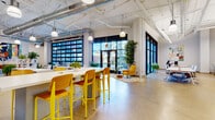 Located at 470 W Broad St in Columbus, Ohio, this single-floor coworking space combines industrial design with warm, contemporary interiors. Soaring ceilings with exposed ductwork and structural elements give the space an open, loft-like character, while polished concrete floors and white walls create a clean, professional backdrop throughout.

The main work area is expansive and bathed in natural light, thanks to floor-to-ceiling black-framed windows and large glass garage-style doors that open the space to street views. Flexible workstations with modern chairs are arranged throughout, accommodating both individual focus work and collaborative efforts. A dedicated lounge area features bold yellow armchairs and a patterned area rug, offering a relaxed setting for informal meetings.

A welcoming reception area greets visitors near the entrance, complete with a geometric chandelier, leather seating, and lush greenery. The coworking lounge displays pegboard accent walls adorned with trailing plants, shelving, and curated décor, adding warmth and personality. Private enclosed office pods and glass-partitioned rooms provide quieter work options, while a fully equipped kitchenette offers a coffee station, microwave, dishwasher, and open shelving stocked with snacks and supplies. Colorful original artwork is displayed throughout, enhancing the creative, community-centered atmosphere of this dynamic workspace.