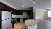 Located at 1211 Gray Birch Way in Knoxville, Tennessee, this single-floor residence offers a thoughtfully designed layout centered around a modern, open-concept living space. The kitchen is a standout feature, showcasing rich dark espresso cabinetry that extends floor to ceiling, providing generous storage throughout. Speckled granite countertops run along the perimeter and continue onto a curved central island, offering additional prep space and a natural gathering point. Stainless steel appliances — including a range, built-in microwave, dishwasher, and refrigerator — complement the contemporary aesthetic. Warm-toned hardwood flooring flows seamlessly from the kitchen into the adjacent living area, creating a cohesive and inviting atmosphere. Overhead, a track lighting system with adjustable fixtures ensures the kitchen is well-lit and functional. The open sightlines between the kitchen and living area lend the space a spacious, airy quality, ideal for everyday living and entertaining. Neutral wall tones throughout contribute to a clean, versatile backdrop that suits a variety of interior styles. The overall design balances practicality with modern style, making this Knoxville home a well-appointed and comfortable residence.