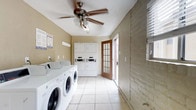 This shared laundry facility is a practical, single-floor space designed to accommodate the needs of multiple users efficiently. The room features a row of front-loading washing machines along one wall, complemented by stacked washer-dryer units positioned near the back, providing ample laundry capacity. The floor is finished with light-toned ceramic tiles that are easy to maintain and contribute to a clean, bright atmosphere. The walls display a warm beige tone, and one side showcases exposed painted brick, adding a utilitarian character to the space. A ceiling fan with an integrated light fixture is centrally mounted, ensuring steady air circulation throughout the room. Natural light filters in through a glass-paneled wooden door that leads to the exterior, as well as through a window fitted with horizontal blinds, keeping the space feeling open and well-ventilated. Informational signage is posted on the wall, providing users with guidelines and instructions for proper machine use. The layout is straightforward and functional, with clear pathways between machines for ease of use. Overall, the facility is a clean, organized, and well-maintained space suited for communal laundry needs.