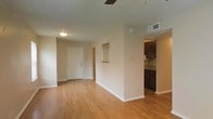 This single-floor residence offers a straightforward, functional layout designed for comfortable everyday living. The main living area is finished with warm hardwood-style laminate flooring that extends throughout, lending a cohesive and clean aesthetic to the space. Neutral beige walls provide a versatile backdrop, while a ceiling fan overhead aids in air circulation. A flush-mount light fixture near the entryway provides additional illumination.

The front entry features a solid white door, and a small recessed niche is built into the dividing wall, offering a decorative display space. A textured ceiling adds subtle dimension to the interior.

Adjacent to the living area, the kitchen is partially visible and features dark espresso-toned cabinetry paired with what appears to be a darker countertop surface. The cabinetry provides ample storage, and the space is compact yet functional. HVAC vents are integrated into the ceiling, ensuring climate control throughout. White baseboards frame each room, adding a clean finishing detail. Overall, the space maintains a simple, well-maintained character suitable for a variety of lifestyles.