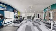 Located in Noblesville, Indiana, this fitness center offers a thoughtfully designed exercise space across a single floor. The room is finished with a bold, contemporary aesthetic, featuring teal, lime green, and white accent walls paired with a textured brick-style wall panel that adds visual depth near the strength training area. A geometric, multi-toned gray carpet tile pattern runs throughout the floor, contributing to the modern and energetic atmosphere.

The cardio zone is well-appointed with a row of TRUE brand treadmills and elliptical machines, all positioned near large windows that invite natural light and views of the surrounding exterior. Wall-mounted flat-screen televisions keep users entertained during workouts. A ceiling fan with recessed lighting above ensures comfortable air circulation throughout the space.

The strength training section includes a multi-station cable machine, a dedicated dumbbell rack stocked with a full range of colorful weights, an adjustable workout bench, and a punching bag mounted along the mirrored wall. The mirrored section expands the sense of space and provides functional visibility during strength exercises.

A built-in nook with a bench and hooks offers a convenient area for storing personal items. Multiple windows along the perimeter flood the room with daylight, creating an open and motivating environment well-suited for a comprehensive fitness routine.