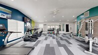 Located in Noblesville, Indiana, this fitness center offers a thoughtfully designed exercise space across a single floor. The room is finished with a bold, contemporary aesthetic, featuring teal, lime green, and white accent walls paired with a textured brick-style wall panel that adds visual depth near the strength training area. A geometric, multi-toned gray carpet tile pattern runs throughout the floor, contributing to the modern and energetic atmosphere.

The cardio zone is well-appointed with a row of TRUE brand treadmills and elliptical machines, all positioned near large windows that invite natural light and views of the surrounding exterior. Wall-mounted flat-screen televisions keep users entertained during workouts. A ceiling fan with recessed lighting above ensures comfortable air circulation throughout the space.

The strength training section includes a multi-station cable machine, a dedicated dumbbell rack stocked with a full range of colorful weights, an adjustable workout bench, and a punching bag mounted along the mirrored wall. The mirrored section expands the sense of space and provides functional visibility during strength exercises.

A built-in nook with a bench and hooks offers a convenient area for storing personal items. Multiple windows along the perimeter flood the room with daylight, creating an open and motivating environment well-suited for a comprehensive fitness routine.