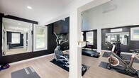 Located at 392 State St in North Haven, CT, this dedicated exercise room delivers an impressive fitness experience within a thoughtfully designed single-floor space. The room's most striking architectural feature is its dramatic vaulted ceiling, which soars to a considerable height and is accented by a bold, geometric black accent wall that rises to meet the roofline. A ceiling fan and a skylight window at the peak of the vault flood the space with natural light and promote airflow, creating a bright and energizing atmosphere.

The interior design follows a clean black-and-white palette, with deep charcoal walls contrasting against crisp white trim, columns, and ceiling. Light wood-toned flooring runs throughout, lending warmth and a polished finish to the overall aesthetic. Multiple windows along the walls further enhance the natural light and provide ventilation.

The fitness area is well-appointed with a variety of cardio equipment, including treadmills, an elliptical, a rowing machine, and a stationary spin bike. A weight bench and a rack of dumbbells round out the strength training offerings. A wall-mounted flat-screen television is positioned for convenient viewing during workouts. Separate restroom facilities are accessible directly from the main floor. The space is both functional and visually refined, making it an exceptional dedicated fitness facility.