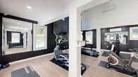 Located at 392 State St in North Haven, CT, this dedicated exercise room delivers an impressive fitness experience within a thoughtfully designed single-floor space. The room's most striking architectural feature is its dramatic vaulted ceiling, which soars to a considerable height and is accented by a bold, geometric black accent wall that rises to meet the roofline. A ceiling fan and a skylight window at the peak of the vault flood the space with natural light and promote airflow, creating a bright and energizing atmosphere.

The interior design follows a clean black-and-white palette, with deep charcoal walls contrasting against crisp white trim, columns, and ceiling. Light wood-toned flooring runs throughout, lending warmth and a polished finish to the overall aesthetic. Multiple windows along the walls further enhance the natural light and provide ventilation.

The fitness area is well-appointed with a variety of cardio equipment, including treadmills, an elliptical, a rowing machine, and a stationary spin bike. A weight bench and a rack of dumbbells round out the strength training offerings. A wall-mounted flat-screen television is positioned for convenient viewing during workouts. Separate restroom facilities are accessible directly from the main floor. The space is both functional and visually refined, making it an exceptional dedicated fitness facility.