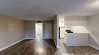 Located at 1049 Brentwood Dr in Daytona Beach, Florida, this single-floor unit offers an efficient and functional layout designed to maximize every square foot. The combined kitchen and living area serves as the heart of the space, featuring durable wood-look flooring in a rich dark tone that runs throughout the unit, complemented by neutral beige walls and clean white baseboards. The living area receives generous natural light through a large front-facing window with blinds, creating a bright and welcoming atmosphere. A white front door with a classic panel design provides a tidy entry point directly into this open area.

The kitchen is well-appointed with white cabinetry above and below the counters, a stainless steel double-basin sink, light-toned countertops, and a black electric range, offering practical functionality in a compact galley-style configuration. A peninsula with beadboard paneling visually separates the kitchen from the living space while maintaining an open feel, with overhead cabinetry providing additional storage.

The bathroom features a full-size soaking tub with a shower, floor-to-ceiling white square tile surround, chrome fixtures, and a frosted glass window that allows light while maintaining privacy. A connecting hallway ties the rooms together, completing this straightforward and efficient floor plan.