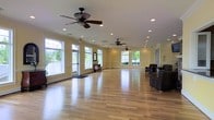 This spacious single-floor interior offers a welcoming and versatile environment well-suited for a variety of uses, from community gatherings to fitness classes or social events. The open floor plan is anchored by warm hardwood flooring that extends throughout the entire space, lending a clean and polished aesthetic. Pale yellow walls contribute to the room's light, airy atmosphere, complementing the natural light that pours in through multiple large windows positioned along the perimeter. These windows frame pleasant outdoor views of lush greenery, enhancing the connection between interior and exterior.

The ceiling features multiple ceiling fans with integrated lighting, supported by recessed lighting throughout that ensures the space is well-illuminated at any time of day. Subtle architectural details, including arched doorways and white trim work, add a touch of classic charm to the otherwise contemporary design. A set of glass-paneled doors provides direct outdoor access, further improving the flow of natural light and ventilation. A small seating area with dark upholstered chairs is tucked along one wall, offering a comfortable resting spot. Overall, the space is functional, bright, and thoughtfully designed.