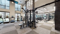This fitness center is designed to deliver a premium workout experience within a thoughtfully appointed, single-level space that benefits from dramatically tall, double-height ceilings. Large floor-to-ceiling windows along the perimeter flood the interior with natural light while offering views of the surrounding building exterior, creating an open and energizing atmosphere.

The cardio zone is lined with multiple treadmills and elliptical trainers, each equipped with individual screens for entertainment. A dedicated strength training area features a full Matrix functional trainer system, a comprehensive dumbbell rack, benches, and additional cable equipment, providing a thorough range of resistance training options. Wall-mounted televisions are positioned throughout the space, ensuring visibility from virtually every workout station.

The interior design incorporates geometric accent wall panels, sleek pendant lighting, and a two-tone modular carpet tile floor that adds visual texture. Full-length mirrors extend the perceived depth of the strength area, while dark structural columns provide industrial contrast against the bright white walls. A glass-paneled entrance with double doors connects the fitness center to adjacent building amenities, completing the cohesive, modern aesthetic.