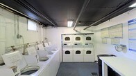 Located at 130 Aberdeen Red in Matawan, New Jersey, this single-level shared laundry facility is designed to serve the practical needs of its users with efficiency and ease. The room is laid out with a row of top-loading washing machines along one side, providing ample capacity for multiple loads at once. Opposite, a bank of stacked commercial-grade dryers lines the wall, maximizing the use of vertical space and offering substantial drying capacity. The interior features white walls that contribute to a clean, utilitarian aesthetic, while overhead fluorescent lighting ensures the space is well-lit throughout. Exposed pipes and ductwork run along the dark-painted ceiling, reflecting the functional, no-frills character of the room. A small folding or sorting surface is available near the entrance, providing a convenient area to manage laundry. Informational notices are posted on the walls, keeping users informed of guidelines and procedures. The flooring is a durable, low-maintenance surface suited to the demands of a high-use laundry environment. Overall, this facility is straightforward and practical, offering everything needed for an efficient laundry experience in a shared-use setting.
