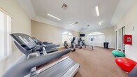 Located at 1900 Bella Vista Way in Port St. Lucie, Florida, this fitness center offers a well-equipped, single-floor workout space designed to support a variety of exercise routines. The room features warm beige walls, textured carpet flooring, and elevated ceilings that contribute to an open, airy atmosphere. Distinctive arched windows allow ample natural light to fill the space, complemented by overhead fluorescent lighting and a ceiling fan for climate comfort. The cardio section is well-stocked with multiple treadmills and elliptical cross-trainers, providing options for users of varying fitness levels. A strength training area includes a weight bench, a cable/multi-use exercise station, and a rack of dumbbells, with a floor-to-ceiling mirror along one wall to assist with form and technique. Stability balls and exercise mats are also available for flexibility and core training. A wall-mounted flat-screen television adds entertainment during workouts. Safety features include a fire extinguisher and emergency exit lighting. A bulletin board near the entrance provides informational postings, and a glass door offers direct access, likely to an outdoor amenity area. The space is thoughtfully arranged to maximize usability within its compact footprint.