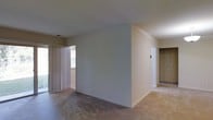 This single-floor residence offers a clean, straightforward layout designed for comfortable everyday living. The main living area is finished with wall-to-wall carpeting in a neutral tone, complementing the crisp white walls that create a bright, versatile backdrop. A large window fitted with floor-to-ceiling vertical drapes allows generous natural light to filter into the space, contributing to an open and airy atmosphere. A central partial wall divides the living area, providing a degree of spatial definition while maintaining an open-plan feel throughout. A hallway visible from the main living area leads to additional rooms, suggesting a functional and efficient floor plan. A closet opening is visible along the far wall, offering practical built-in storage. The overall ambiance is calm and neutral, providing a blank canvas that can be easily adapted to a variety of interior styles and furnishing arrangements. The single-floor configuration ensures convenient, step-free access throughout the entire unit, making it a practical and accessible living space.