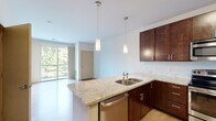 This single-floor residence showcases a thoughtfully designed open layout that seamlessly connects the kitchen and living areas. The kitchen is a standout feature, equipped with rich dark-wood cabinetry, granite countertops, and a peninsula with a double stainless steel sink, ideal for casual dining or meal preparation. Stainless steel appliances — including a range, built-in microwave, dishwasher, and refrigerator — complement the modern aesthetic. Pendant lighting above the peninsula adds a stylish focal point.

The living area is bright and airy, with light wood-style flooring and a large sliding glass door that opens to a balcony, framing lush green tree views and filling the space with natural light. The bedroom features soft carpeting, a spacious closet with built-in shelving, and direct access to the rest of the unit. The bathroom is finished with large-format dark tile flooring, a dark-wood vanity with a white countertop, a sizable mirror, and a well-lit vanity fixture. Throughout the unit, clean white walls and consistent modern finishes create a cohesive, contemporary ambiance.