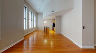 Located on the 5th floor at 911 Locust St in downtown St. Louis, this unit combines modern finishes with an open, airy design. The main living area impresses with soaring vaulted ceilings, warm hardwood flooring, and a wall of tall windows that flood the space with natural light. A graceful curved architectural detail defines the transition between the living area and the kitchen, adding a distinctive design element. The kitchen is well-appointed with honey-toned cabinetry, dark granite countertops, stainless steel appliances including a side-by-side refrigerator and dishwasher, and a central island with a bar-height overhang — ideal for casual dining. Pendant lighting above the island and kitchen adds a refined touch. The bedroom features plush carpeting, a ceiling fan, and windows overlooking neighboring architecture. A second room with hardwood floors and a ceiling fan offers flexible use as an additional bedroom or office. The bathroom is finished with marble tile surrounding a deep soaking tub, tile flooring, and built-in wood cabinetry with granite countertops. A dedicated laundry closet with a stacked Electrolux washer and dryer adds everyday convenience. Throughout, the unit maintains a consistent, polished aesthetic with neutral tones and quality materials.