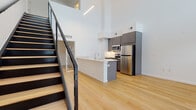 This contemporary two-floor loft-style space is defined by its dramatic double-height ceilings and clean, modern aesthetic. The main level showcases an open-concept layout with light hardwood flooring that flows seamlessly throughout. The kitchen is thoughtfully designed with charcoal-toned cabinetry, a full suite of stainless steel appliances including a refrigerator, microwave, and range, and a peninsula with a built-in sink and light countertops — ideal for both cooking and casual dining.

A standout architectural feature is the sleek floating staircase with a black steel frame and cable railings, leading to the upper level. The second floor is bathed in natural light from large windows and opens to a bright, versatile area suitable for a bedroom or living space, complete with the same warm hardwood flooring and crisp white walls.

Two full bathrooms are finished consistently with neutral tile flooring, soaker tubs with shower combinations, and dark-toned vanities topped with light countertops and large mirrors. The overall ambiance throughout the space is bright, airy, and minimalist, with a cohesive modern design that balances clean lines with functional, well-appointed details across both levels.