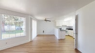 This single-floor unit presents a clean, minimalist aesthetic throughout, defined by crisp white walls, light hardwood-style flooring, and an open layout that promotes a sense of spaciousness. The living area serves as a welcoming focal point, centered around a dark-surround fireplace that provides visual contrast against the otherwise bright, neutral palette. The room is generously proportioned, offering ample flexibility for furniture arrangement.

The living space flows directly into the kitchen and dining area, creating a cohesive, open-plan environment. Large windows with vertical blinds admit abundant natural light and offer views of surrounding greenery. A ceiling fan overhead adds both function and a finishing detail to the room.

The kitchen is efficiently designed with white shaker-style cabinetry extending to the upper walls, dark countertops, and a peninsula that separates the cooking area from the living space. White appliances — including a range, refrigerator, and dishwasher — are included. Flush overhead lighting keeps the workspace well-illuminated.

The bathroom features a full tub and shower combination with clean white surrounds and chrome fixtures, maintaining the unit's consistent, understated aesthetic throughout.