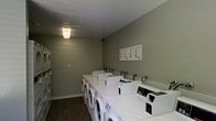 This communal laundry facility is a practical, single-floor space designed to accommodate multiple users efficiently. The room features a row of front-loading washing machines neatly aligned along the wall, complemented by additional stacked laundry units that maximize the available space. The machines are uniformly white, lending the room a clean and orderly appearance.

The walls are finished in a neutral sage green tone, creating a calm and utilitarian ambiance. Dark hardwood-style flooring runs throughout, providing a durable and easy-to-maintain surface that contrasts neatly with the white appliances. Overhead, a flush-mounted light fixture provides steady, even illumination across the room.

An instructional notice board is mounted on the far wall, offering users clear guidance on operating the machines. The layout is thoughtfully organized, allowing for comfortable movement between machines and easy loading and unloading access. A ceiling panel, likely housing ventilation components, ensures proper airflow within the space.

Overall, this laundry room is a well-maintained, efficiently designed facility that prioritizes function and ease of use, making it a reliable shared amenity for its users.