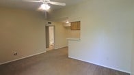 This single-story space in Texarkana offers a straightforward, functional layout centered around an open living area finished with wall-to-wall carpeting in a neutral tone. The room is equipped with a ceiling fan with integrated lighting, providing both airflow and illumination throughout the space. A half-wall partition separates the living area from the kitchen, featuring a pass-through opening accented by a wooden-paneled detail above, allowing for easy communication between the two areas. The kitchen pass-through adds a sense of openness while maintaining a defined boundary between the spaces. The neutral-toned walls throughout create a clean, understated backdrop. A doorway visible from the main living area leads directly to a bathroom, hinting at a compact and efficient floor plan. Recessed ceiling vents suggest a central HVAC system for climate control. The overall ambiance is simple and practical, with a layout designed for ease of use and straightforward livability.