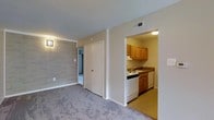 Located at 411 East Gibbsboro Road in Lindenwold, New Jersey, this single-floor residential unit offers a practical and comfortable layout designed for everyday living. The living area features soft, neutral-toned carpeting and a distinctive stone-textured accent wall that adds character to the space. A wall-mounted light fixture complements the understated aesthetic, while a ceiling vent ensures climate-controlled comfort throughout. Adjacent to the living area, a closet provides convenient storage, and an open passageway leads directly into the kitchen. The kitchen is outfitted with warm-toned wooden cabinetry offering ample storage, dark granite-style countertops, and white appliances including a range and dishwasher. A stainless steel sink sits beneath the upper cabinets, making the space both efficient and functional. The kitchen's tile flooring contrasts neatly with the carpeted living area, visually defining each zone. A hallway visible from the main living space suggests additional rooms beyond, contributing to an organized and connected floor plan. Overall, the unit presents a clean, move-in ready environment with a straightforward design that prioritizes functionality and comfort.