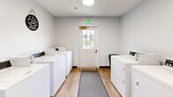 Located at 4211 W Potomac Dr in Greenfield, Indiana, this shared laundry facility is a compact, efficiently designed single-room space. The room is finished with clean white walls and warm wood-look luxury vinyl plank flooring, accented by dark baseboards that lend a tidy, polished appearance throughout.

Multiple commercial-grade top-load washing machines and dryers are positioned along both sides of the room, providing ample laundry capacity. A long folding table with accompanying seating runs along one wall, offering a convenient surface for sorting and folding laundry. Gray runner rugs placed along the center of the floor add a practical touch, protecting the flooring in high-traffic areas.

The room is well-lit by flush-mounted ceiling lights, and a windowed entry door at one end allows natural light to filter in while also providing a view to the outdoors. Posted signage and a bulletin board on the wall communicate laundry schedules and usage guidelines for occupants. A fire extinguisher is prominently mounted near the entry, underscoring the room's attention to safety. Overall, the space is clean, functional, and thoughtfully organized to serve the needs of its users efficiently.