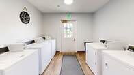 Located at 4211 W Potomac Dr in Greenfield, Indiana, this shared laundry facility is a compact, efficiently designed single-room space. The room is finished with clean white walls and warm wood-look luxury vinyl plank flooring, accented by dark baseboards that lend a tidy, polished appearance throughout.

Multiple commercial-grade top-load washing machines and dryers are positioned along both sides of the room, providing ample laundry capacity. A long folding table with accompanying seating runs along one wall, offering a convenient surface for sorting and folding laundry. Gray runner rugs placed along the center of the floor add a practical touch, protecting the flooring in high-traffic areas.

The room is well-lit by flush-mounted ceiling lights, and a windowed entry door at one end allows natural light to filter in while also providing a view to the outdoors. Posted signage and a bulletin board on the wall communicate laundry schedules and usage guidelines for occupants. A fire extinguisher is prominently mounted near the entry, underscoring the room's attention to safety. Overall, the space is clean, functional, and thoughtfully organized to serve the needs of its users efficiently.