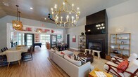 Located at 1818 Wards Ferry Ridge in Lynchburg, Virginia, this single-story clubhouse-style space is designed to accommodate a variety of social and recreational activities within a thoughtfully curated interior. The open floor plan flows seamlessly across light hardwood flooring, connecting multiple functional zones while maintaining a cohesive, contemporary aesthetic throughout.

The central lounge area is a striking focal point, anchored by a tall dark-surround fireplace with a wall-mounted television above it, flanked by gold open shelving. A large sectional sofa with bold patterned accent pillows, a tufted leather ottoman, and mixed seating arrangements create an inviting gathering space beneath a statement gold candelabra chandelier that rises to soaring vaulted ceilings.

Adjacent dining areas include both a large communal table with upholstered chairs illuminated by geometric gold pendant lights and a smaller round bistro-style table for informal seating. French glass doors connect the main lounge to a dedicated game room featuring a pool table, bold ikat-patterned wallcovering, and a media wall. A kitchenette with granite countertops, dark cabinetry, and vibrant terracotta-painted walls adds functional amenity. Natural light flows throughout from multiple windows, enhancing the welcoming ambiance.