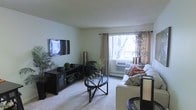 This single-floor space showcases a thoughtfully arranged living room that serves as its central feature. The room is finished with neutral, light-toned walls and a textured ceiling, creating a calm and understated atmosphere. Soft, wall-to-wall carpeting adds warmth underfoot and contributes to the overall coziness of the space.

A large window fitted with grommet-style curtains in a warm beige tone allows natural light to flow into the room, offering a view of mature trees outside. A wall-mounted air conditioning unit beneath the window provides climate control. The living area is anchored by a light-colored sofa accented with patterned and solid throw pillows, paired with a stylish round glass-top coffee table that adds a modern touch.

A dark-toned media console along the opposite wall houses a flat-screen television mounted above it, complemented by decorative plants and accessories that bring life and greenery to the space. A sleek floor lamp adds additional lighting. The overall ambiance is tidy, comfortable, and inviting, with a balanced mix of functional and decorative elements.