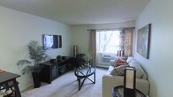 This single-floor space showcases a thoughtfully arranged living room that serves as its central feature. The room is finished with neutral, light-toned walls and a textured ceiling, creating a calm and understated atmosphere. Soft, wall-to-wall carpeting adds warmth underfoot and contributes to the overall coziness of the space.

A large window fitted with grommet-style curtains in a warm beige tone allows natural light to flow into the room, offering a view of mature trees outside. A wall-mounted air conditioning unit beneath the window provides climate control. The living area is anchored by a light-colored sofa accented with patterned and solid throw pillows, paired with a stylish round glass-top coffee table that adds a modern touch.

A dark-toned media console along the opposite wall houses a flat-screen television mounted above it, complemented by decorative plants and accessories that bring life and greenery to the space. A sleek floor lamp adds additional lighting. The overall ambiance is tidy, comfortable, and inviting, with a balanced mix of functional and decorative elements.