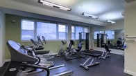 The fitness center at 239 W Johnson Street in Philadelphia's Germantown neighborhood offers a comprehensive workout facility on a single level. The space is outfitted with a variety of cardio equipment, including treadmills and stationary recumbent and upright bikes, providing options for users at different fitness levels. A cable machine and weight bench round out the strength training offerings, making the facility suitable for full-body workouts. Large windows line one wall, allowing natural light to fill the space and providing views of the surrounding neighborhood. Overhead, bright fluorescent strip lighting ensures consistent illumination throughout. A wall-length mirror on one side of the room enhances the sense of space while allowing users to monitor their form during exercise. The flooring consists of durable dark rubber matting, a practical choice designed to withstand heavy equipment and high foot traffic. The walls are painted in a two-tone scheme of white and olive green, creating a clean, functional environment. Overall, the fitness center is designed to be a practical and accessible amenity, balancing essential equipment with a comfortable, well-lit atmosphere.