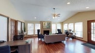 This single-floor residence offers a thoughtfully designed layout centered around a bright and spacious open living area. The room immediately draws attention with its vaulted ceilings, which lend an airy, expansive feel to the interior. A textured ceiling adds subtle architectural character throughout.

Rich hardwood flooring flows across the main living space, providing warmth and a polished aesthetic. The focal point of the room is a classic brick fireplace, framed by a bold piece of abstract artwork that adds a contemporary touch to the otherwise traditional design. Multiple windows dressed with horizontal blinds line the walls, flooding the space with generous natural light while offering privacy when needed.

A ceiling fan with a brass fixture is centrally mounted, providing both functional airflow and a timeless decorative element. Recessed lighting complements the natural light, ensuring the space is well-lit at any hour. The living area is versatile, accommodating a comfortable seating arrangement as well as a dedicated workspace, complete with a desk and computer setup tucked neatly into one corner. A decorative console table along one wall adds additional style and storage. Overall, the space strikes a balance between comfort and functionality, making it well-suited for both relaxing and working from home.