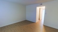 This two-floor space is characterized by its clean, understated design and practical layout. The interior showcases warm hardwood-style flooring that runs throughout, complementing the crisp white walls and creating a bright, welcoming atmosphere. The main living area is open and well-proportioned, with smooth white walls, white baseboards, and a textured ceiling that adds subtle dimension overhead. A ceiling vent indicates a central HVAC system for year-round climate control. An open passageway connects the main living area to an adjacent hallway, which leads further into the space. Natural light filters in through what appears to be a window dressed with white vertical blinds, brightening the corridor and contributing to the overall airy feel of the interior. The neutral color scheme throughout provides a versatile backdrop suitable for a variety of furnishing styles. The layout suggests a logical flow between living areas, with the hallway offering access to additional rooms across the two floors. Overall, the space presents a tidy, move-in-ready environment with consistent finishes and a straightforward, functional design.
