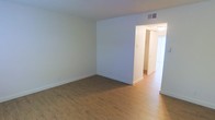 This two-floor space is characterized by its clean, understated design and practical layout. The interior showcases warm hardwood-style flooring that runs throughout, complementing the crisp white walls and creating a bright, welcoming atmosphere. The main living area is open and well-proportioned, with smooth white walls, white baseboards, and a textured ceiling that adds subtle dimension overhead. A ceiling vent indicates a central HVAC system for year-round climate control. An open passageway connects the main living area to an adjacent hallway, which leads further into the space. Natural light filters in through what appears to be a window dressed with white vertical blinds, brightening the corridor and contributing to the overall airy feel of the interior. The neutral color scheme throughout provides a versatile backdrop suitable for a variety of furnishing styles. The layout suggests a logical flow between living areas, with the hallway offering access to additional rooms across the two floors. Overall, the space presents a tidy, move-in-ready environment with consistent finishes and a straightforward, functional design.