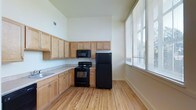 This single-floor space centers around a well-appointed kitchen that combines practicality with a clean, understated aesthetic. The kitchen features light maple wood cabinetry arranged in an L-shaped configuration, offering generous upper and lower storage throughout. Countertops provide ample workspace, complementing the full suite of black appliances that includes a refrigerator, electric range, and over-the-range microwave. Light hardwood flooring runs throughout, lending warmth and a cohesive visual flow to the space. The elevated ceiling height contributes to an open, airy atmosphere, making the kitchen feel notably spacious. A large window along the side wall allows substantial natural light to fill the room, enhancing the bright, welcoming environment. The neutral white walls provide a clean backdrop that keeps the space feeling fresh and versatile. The overall design is straightforward and functional, well-suited for everyday use, with a layout that promotes ease of movement and accessibility to all key kitchen areas.