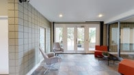 This single-floor space presents a well-appointed lobby or reception area that blends modern design with functional elegance. The flooring is composed of natural stone tiles in varied earth tones, lending a grounded, sophisticated feel to the room. A prominent feature wall clad in square ceramic or stone tiles in a neutral grid pattern adds architectural texture and visual interest, complemented by a frosted glass panel that introduces a refined decorative element.

The seating arrangement includes a pair of contemporary lounge chairs — one in soft blush and another in a complementary neutral tone — alongside a bold orange accent sofa, creating a welcoming and stylistically dynamic conversation area. Recessed ceiling lighting provides consistent, even illumination throughout the space.

Large white-framed glass entry doors span much of the far wall, flooding the interior with natural light while offering unobstructed views of lush palm trees and landscaped outdoor surroundings. Dark fabric curtain panels frame the doorway, adding contrast and a touch of warmth. The overall ambiance is clean, contemporary, and inviting, reflecting a well-maintained and thoughtfully designed public or communal entrance space.