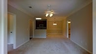 This single-floor space showcases an open-concept design that connects the living area to the kitchen, creating a functional and cohesive floor plan. The living area is finished with plush wall-to-wall carpeting and features crown molding along the ceiling, adding a traditional architectural detail. A ceiling fan with a light fixture is centrally positioned, providing both ventilation and ambient lighting.

The kitchen is partially open to the living space via a raised bar-style counter, offering a natural divide while maintaining visual connectivity. Dark cabinetry lines the kitchen walls, complemented by a stainless steel microwave and a cooktop range. An overhead fluorescent light illuminates the kitchen workspace efficiently.

A pendant chandelier with warm-toned bulbs is suspended in what appears to be a designated dining area, adding a decorative focal point to that zone. Doorways visible on both sides of the living area lead to additional rooms, with one appearing to provide access to a bathroom or secondary space, hinting at a practical and well-organized layout throughout the home.