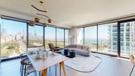 Located at 1210 S Indiana Ave in Chicago's vibrant South Loop neighborhood, this thoughtfully designed residence offers an exceptional living experience defined by modern finishes and breathtaking panoramic views. The open-concept living and dining area is anchored by expansive floor-to-ceiling corner windows that frame sweeping vistas of Lake Michigan, Grant Park, and the Chicago skyline, flooding the space with natural light throughout the day. A sculptural pendant light fixture with amber globe accents adds warmth to the dining area, which is complemented by a large-scale abstract art piece. The kitchen features sleek, light wood cabinetry, stainless steel appliances including a gas range and French door refrigerator, and a generous island with a clean-lined countertop. The bedroom includes a wall-mounted display, a dedicated workspace, and direct access to a closet and a well-appointed en-suite. The bathroom is finished with clean white tile surrounding a soaking tub and shower combination. An in-unit stacked washer and dryer provides added convenience. A hallway gallery wall showcasing a dramatic nature-inspired photographic triptych adds an artistic focal point, enhancing the overall sophisticated and curated ambiance of the space.