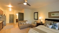 This single-floor residence offers a well-organized layout with functional design throughout. The galley-style kitchen is appointed with rich dark wood cabinetry, light countertops, and a full suite of stainless steel appliances, including a refrigerator, microwave, and dishwasher. A double-basin sink sits beneath the cabinetry, and the kitchen flows naturally into an entryway corridor featuring light hardwood-style flooring and flush-mount ceiling lighting. Decorative touches, including tall bamboo accents and lush tropical plants, add a warm, natural element near the kitchen area.

The bedroom is generously proportioned, featuring plush wall-to-wall carpeting and a ceiling fan with integrated lighting to ensure year-round comfort. Warm, neutral tones define the room's aesthetic, complemented by soft ambient lighting. A sitting area with an upholstered chaise and accent chair offers additional comfort and versatility. The bedroom provides direct access to an ensuite bathroom, visible through a charcoal-painted door, which includes vanity cabinetry and mirror. Overall, the space presents a clean, contemporary feel with cohesive finishes throughout.