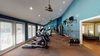 This fitness center in Conroe, Texas offers a comprehensive workout environment designed to accommodate a wide range of exercise needs. The space is defined by striking teal-painted walls, vaulted ceilings with recessed lighting, and industrial-style ceiling fans that contribute to both the aesthetic appeal and ventilation of the room. Durable carpet tile flooring covers the workout area, providing a practical and comfortable surface underfoot. A row of large windows along one wall invites abundant natural light, creating an open and energizing atmosphere. The cardio section is well-stocked with treadmills, elliptical machines, and stationary bikes positioned along the length of the room. A full-wall mirror visually expands the space while allowing users to monitor their form. On the strength training side, the facility includes a multi-station cable machine, a power rack with pull-up bar, a weight bench with free weights and plates, and a heavy punching bag for functional training. Wall-mounted flat-screen televisions are strategically placed throughout for entertainment during workouts. Framed artwork adds a decorative touch to the space. An adjoining room with a glass-paneled door offers additional utility, and a small beverage refrigerator provides convenient refreshment access.