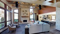 This single-story space is defined by its impressive architectural character and warm, inviting ambiance. The centerpiece of the main lounge is a dramatic floor-to-ceiling stone fireplace, flanked by tall windows that draw in abundant natural light and create a sense of openness. Exposed timber beams and trusses span the soaring ceiling overhead, lending a rustic yet refined aesthetic that is complemented by industrial-style ductwork and a geometric pendant chandelier.

The lounge area is generously proportioned, furnished with a large sectional sofa and accent chairs in complementary earthy tones, arranged around a solid wood coffee table for a comfortable and functional gathering space. Warm terracotta accent walls contrast beautifully with the neutral stone and wood elements throughout the room.

Adjacent to the lounge is a built-in refreshment or café bar area, featuring open shelving and counter space, ideal for serving residents or guests. The flooring throughout is a durable, dark-toned material that grounds the space and ties together the varied design elements. Overall, the space exudes a lodge-inspired warmth with modern touches, making it well-suited as a community lounge or social gathering area.