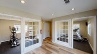 This single-floor fitness center in Savannah, Georgia offers a well-organized layout designed to support a variety of workout routines. The facility is accessed through a bright, open lobby area featuring light wood-style flooring, recessed lighting, and warm neutral-toned walls that create a clean and inviting atmosphere. Large French-style glass-paned doors with white trim divide the lobby from the dedicated exercise rooms, allowing natural light to flow throughout while providing clear sightlines into the workout areas.

The exercise rooms are carpeted in dark, durable flooring appropriate for fitness equipment and feature recessed lighting alongside natural light from windows fitted with blinds. One room is equipped with treadmills, while another houses an elliptical machine and a stationary cycling bike, offering a well-rounded selection of cardio equipment. Both rooms maintain a spacious, uncluttered feel that supports comfortable movement during workouts.

The lobby area also includes built-in white cabinetry with glass-front upper doors and counter space, suggesting storage and convenience amenities for users. A fire extinguisher is visibly mounted in the hallway, reflecting attention to safety standards. The overall design balances functionality with a tidy, welcoming aesthetic suited to a community fitness facility.