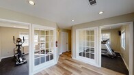 This single-floor fitness center in Savannah, Georgia offers a well-organized layout designed to support a variety of workout routines. The facility is accessed through a bright, open lobby area featuring light wood-style flooring, recessed lighting, and warm neutral-toned walls that create a clean and inviting atmosphere. Large French-style glass-paned doors with white trim divide the lobby from the dedicated exercise rooms, allowing natural light to flow throughout while providing clear sightlines into the workout areas.

The exercise rooms are carpeted in dark, durable flooring appropriate for fitness equipment and feature recessed lighting alongside natural light from windows fitted with blinds. One room is equipped with treadmills, while another houses an elliptical machine and a stationary cycling bike, offering a well-rounded selection of cardio equipment. Both rooms maintain a spacious, uncluttered feel that supports comfortable movement during workouts.

The lobby area also includes built-in white cabinetry with glass-front upper doors and counter space, suggesting storage and convenience amenities for users. A fire extinguisher is visibly mounted in the hallway, reflecting attention to safety standards. The overall design balances functionality with a tidy, welcoming aesthetic suited to a community fitness facility.