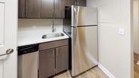 This single-floor unit offers a clean, modern aesthetic throughout, characterized by warm wood-look laminate flooring, neutral beige walls, and crisp white trim and baseboards that create a bright, cohesive atmosphere across all rooms.

The open living area is generously proportioned, providing ample space for a variety of furniture arrangements. Natural light flows in through large windows fitted with horizontal blinds, while a sliding glass door opens directly onto a private balcony, further enhancing the sense of openness. A textured ceiling adds subtle visual interest overhead.

The kitchen is efficiently designed and features dark wood cabinetry, light stone-look countertops, a stainless steel sink, and a full-size stainless steel refrigerator. The layout makes practical use of available space with well-organized upper and lower cabinet storage.

The bedroom is a comfortable size, finished with the same wood-look flooring as the rest of the unit, and includes direct access to an en suite bathroom equipped with a glass-enclosed shower and modern fixtures. A second bathroom features a granite-look countertop with an undermount sink, a wall-mounted mirror, and contemporary chrome hardware. Both bathrooms maintain a clean, minimalist style consistent with the overall design of the space.