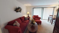 This single-floor residence offers a thoughtfully laid-out living space with a warm and inviting ambiance throughout. The living room serves as the heart of the home, featuring plush carpeting, large windows dressed with vertical blinds that allow ample natural light, and generous space for comfortable seating arrangements. An adjoining sitting or flex area extends the living space further, ideal for reading or relaxing.

The dining area is situated conveniently near the kitchen and features a ceiling fan with light fixture, vertical blinds along a sliding door, and enough room for a full dining table and chairs. The galley-style kitchen is efficiently designed with warm wood cabinetry, dark granite-style countertops, a gas range, refrigerator, and a double sink — with a direct sightline into the dining area, making entertaining easy.

The bedroom is a peaceful retreat with soft carpeting, a large window with vertical blinds, and ample room for a full bedroom suite. A spacious walk-in closet fitted with wire shelving and hanging rods provides excellent storage capacity. The full bathroom features a wood-tone vanity with under-sink storage, a lighted mirror, tile flooring, and a shower-tub combination with a curtain enclosure.