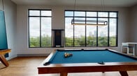 This contemporary single-floor amenity space is thoughtfully designed to offer a variety of recreational and social environments, unified by a bold, modern aesthetic and an abundance of natural light. Floor-to-ceiling windows with black metal frames run throughout the space, framing impressive views of a lush tree canopy and a city skyline beyond.

The game room is a standout area, centered around a full-size billiards table with a teal felt surface beneath a sleek rectangular pendant light. Warm hardwood flooring extends throughout much of the space, complementing the bright, open atmosphere. A shuffleboard table is positioned along a deep teal accent wall adorned with two large abstract artworks, adding a striking visual anchor to the corridor.

The lounge area features vibrant blue sofas, yellow accent chairs, and colorful artwork, all set against expansive windows that invite the outdoors in. Nearby, a media lounge offers a relaxed setting with a large wall-mounted television framed by white brick tile, accompanied by a plush red sectional and bean bag seating beneath a ceiling fan.

The kitchen is well-appointed with two-tone cabinetry, a teal geometric tile backsplash, white countertops, a large island with seating, and warm yellow pendant lighting—ideal for entertaining. Restrooms are conveniently accessible via a connecting hallway, completing this comprehensive and stylishly designed amenity floor.