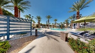 Located at 3140 Saint Rose Pkwy in Henderson, Nevada, this outdoor amenity area delivers a resort-inspired experience designed for relaxation and leisure. The centerpiece of the space is a generously sized swimming pool with a freeform shape, complemented by an adjacent spa, both set against a backdrop of towering mature palm trees that evoke a classic desert oasis atmosphere.

The expansive pool deck is finished in smooth concrete and offers ample room for lounging, with numerous chaise lounge chairs positioned throughout, many shaded by vibrant blue and green market umbrellas. Decorative concrete planters and desert-adapted landscaping add visual interest while reinforcing the sun-drenched Nevada aesthetic.

A standout feature of the space is the modern pergola-style cabana structure, constructed with a bold blue steel frame and horizontal slatted panels that cast dramatic striped shadows across the seating area below. This covered lounge is furnished with navy blue sofas, accent chairs with green striped cushions, and sleek silver side tables, creating an inviting social gathering spot with partial shade and open-air ventilation.

The adjacent building features a contemporary architectural design with large glass windows and clean stucco facades. Stylish black lamp posts are distributed throughout the courtyard, providing evening illumination. The overall ambiance is bright, modern, and resort-caliber, ideal for outdoor recreation in the Henderson, NV climate.