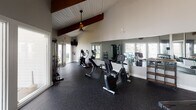Located at 1800 William Kennerty Dr in Charleston, South Carolina, this single-floor fitness center is thoughtfully designed to support a comprehensive workout experience. The space is defined by its striking vaulted ceiling adorned with rich, dark-stained exposed wood beams set against white shiplap, creating a bright and welcoming architectural character that feels both rustic and modern.

Natural light pours in through multiple large windows and glass-panel doors positioned along the perimeter, offering views of the surrounding neighborhood and keeping the space feeling open and airy. Durable rubber flooring in a speckled finish covers the entire workout area, providing comfort and safety underfoot.

The fitness floor is well-equipped with a variety of cardio machines, including stationary bikes, an elliptical trainer, and additional cycling equipment, alongside strength training stations such as a multi-function cable machine, an adjustable weight bench, and a full dumbbell rack with weights of varying sizes. A medicine ball and stability ball round out the functional fitness offerings.

Full-length mirrors line one wall, visually expanding the space and supporting proper form during exercise. Pendant lights and ceiling fans hang from the vaulted ceiling, ensuring comfortable airflow and adequate lighting throughout. Large glass partition walls connect the fitness center to an adjacent community space, further enhancing the open, integrated feel of the facility.
