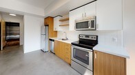 This single-floor unit showcases a clean, contemporary design characterized by polished concrete floors, crisp white walls, and an open, airy layout. The main living area is generously proportioned, benefiting from tall, narrow windows that invite abundant natural light, casting bright patches across the smooth floor. A structural concrete column adds an industrial touch to the open space, while a wall-mounted heating unit ensures year-round comfort.

The kitchen is thoughtfully designed, combining warm wood-grain cabinetry with white upper cabinets and open shelving for a balanced, modern aesthetic. White subway tile backsplash and light countertops complement the full suite of stainless steel appliances, including a refrigerator, electric range, over-the-range microwave, and dishwasher. A single-basin sink with a sleek faucet completes the functional workspace.

The bedroom is a calm, well-lit room featuring polished concrete floors, white walls, and a pair of built-in closets providing generous storage. The bathroom is cleanly finished with a wood-grain vanity, white countertop, a large mirror, and a combined tub and shower, maintaining the unit's consistent modern palette throughout.
