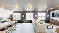 Located in the Fishtown neighborhood of Philadelphia, this contemporary community lounge at 1901 E York St offers a well-appointed, single-floor space designed for comfort and social interaction. The open layout is anchored by a generously sized living area featuring large, light-filled windows that frame views of the surrounding cityscape. Dark charcoal accent walls contrast elegantly with the bright large-format tile flooring, creating a modern yet inviting atmosphere enhanced by warm globe ceiling fixtures throughout.

The lounge area is thoughtfully furnished with a plush sectional sofa, upholstered accent chairs, and a central coffee table, providing ample seating for relaxation or casual gatherings. A wall-mounted flat-screen television and a wood media console anchor one end of the room, while a glass door opens directly to an outdoor terrace, extending the living space into the open air.

Adjacent to the main lounge, a kitchen bar area features a long counter with bar stool seating, backed by a bold geometric patterned tile accent wall with a playful "Yes Chef" sign. A cozy bistro-style table and leather seating add character to this social zone. The space is further enhanced by a sun-drenched seating nook with floor-to-ceiling sliding glass panels, infusing the interior with abundant natural light and offering seamless indoor-outdoor connectivity.