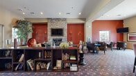 This well-appointed community clubhouse or lounge offers a thoughtfully designed open floor plan across a single level, providing ample space for socializing and relaxation. The interior is anchored by a striking floor-to-ceiling stacked stone fireplace flanked by a wall-mounted flat-screen television, creating a warm and inviting focal point. Rich terracotta-red accent walls contrast beautifully with the cream ceiling, lending the space a bold yet welcoming ambiance. Recessed lighting throughout ensures the area is well-lit, while wall sconces add a layer of atmospheric warmth.

Multiple seating arrangements are distributed across the space, including a cozy lounge area near the fireplace, and separate clusters of striped wingback chairs around round tables for more intimate conversation. A low open bookshelf serves as a room divider, displaying a curated collection of books and decorative objects. The space also features a stylish bar with a dark granite countertop, wood veneer paneling, and pendant lighting with distinctive orange drop shades — ideal for entertaining. Adjacent to the bar is a fully equipped kitchen with dark cabinetry, granite countertops, stainless steel appliances, and tile flooring. Glass entry doors flood the entrance with natural light and provide views to the exterior.