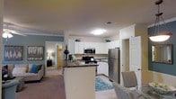 This single-story residence in Morrisville, North Carolina, offers an open-concept layout that connects the kitchen and living areas seamlessly. The kitchen is a functional centerpiece, featuring crisp white cabinetry that extends to the upper walls, providing ample storage space. Dark granite-style countertops offer a contrasting visual appeal, while the breakfast bar peninsula creates a natural divide between the kitchen and the adjoining living space. Stainless steel appliances, including a refrigerator, microwave, and range, lend a modern, polished look to the space.

The flooring transitions from tile in the kitchen to plush carpeting in the living and bedroom areas, adding warmth and comfort underfoot. Soft, neutral wall tones in sage green and beige create a calming atmosphere throughout. Crown molding adds an architectural detail that elevates the overall aesthetic. A doorway visible from the kitchen leads to a carpeted bedroom, suggesting a well-connected floor plan. The layout balances practicality with comfort, making it a well-rounded living space suitable for everyday living in the Morrisville area.
