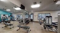 Located at 502 S Freemont Ave in Tampa, FL, this fitness center is a well-appointed, single-floor facility designed to support a full range of workout routines. The space is defined by its open, airy layout, with vaulted ceilings in sections that add a sense of volume and openness throughout.

The facility is divided into distinct workout zones. The cardio area features a lineup of Life Fitness treadmills, elliptical trainers, and a StairMaster, all positioned near large windows that allow natural light to flow in and provide views of the surrounding greenery. A vibrant teal accent wall energizes this section of the space.

The strength training area offers a comprehensive selection of Life Fitness selectorized machines, a multi-function cable machine, and a well-stocked dumbbell rack with a adjustable weight bench. Full-length mirrors line the walls, visually expanding the space while allowing users to monitor their form during workouts.

Overhead, wall-mounted flat-screen televisions are positioned throughout the gym for entertainment during workouts. Ceiling fans and recessed lighting ensure the environment remains comfortable and well-lit. Durable, neutral-toned carpet flooring covers the entire space, providing a practical and clean surface underfoot. A glass-paneled entry door adds a polished, professional finish to the overall aesthetic of this well-maintained fitness facility.